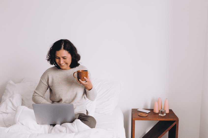 woman sitting on her bed and using her laptop while holding a mug